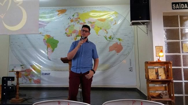 A foto mostra Davi falando em um microfone em frente a um mapa Mundi que ocupa uma parede inteira.
