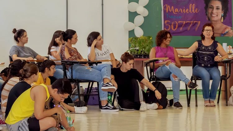 Grupo de mulheres sentadas em roda, algumas em cadeiras e outras no chão. No fundo, uma faixa com a foto de Marielle Franco e o número da sua candidatura nas eleições do Rio de Janeiro, 50777.