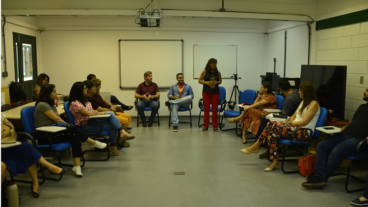Em uma sala de aula, um grupo de 14 pessoas aparece sentado, em círculo. A reitora, professora Cleuza, está em pé, falando. 