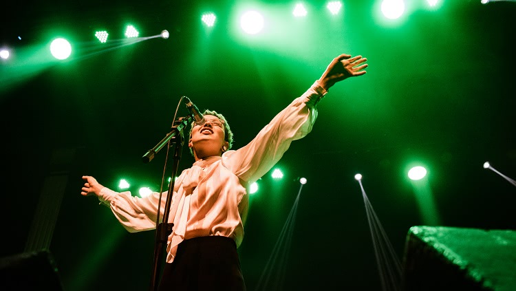 A foto mostra uma cantora em performance num palco. Ela canta ao microfone e está com os braços abertos. Aparece a iluminação do palco em tons de verde.
